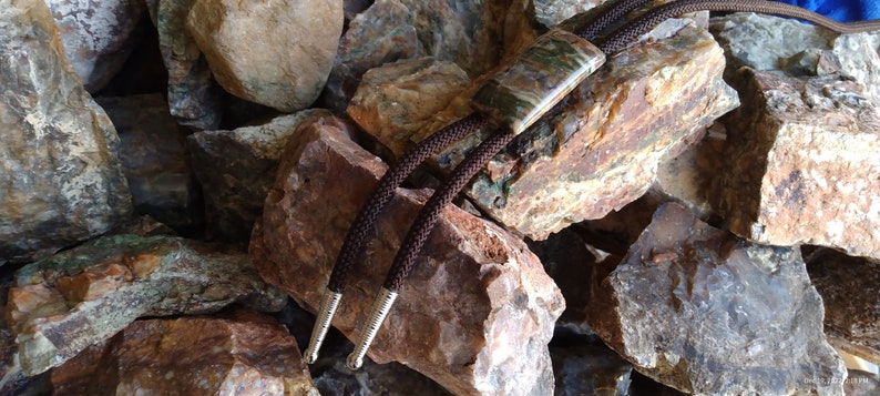 May include: A bolo tie with a rectangular, patterned stone pendant in shades of green, brown, and cream. The tie features a dark brown braided cord with silver-colored tips. The bolo tie rests on a bed of various-sized rocks.