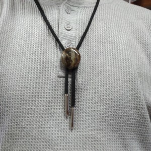 Pu&ograve; includere: Un bolo tie nero con un ciondolo rotondo in pietra marrone e bianca. Il ciondolo ha un bordo in metallo argentato. Il bolo tie &egrave; indossato da una persona che indossa una camicia grigia a nido d'ape.