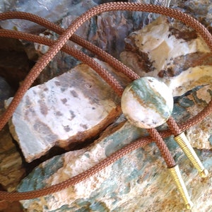 May include: A brown leather bolo tie with a round green and white agate stone pendant. The bolo tie is displayed on a bed of green and white rocks.
