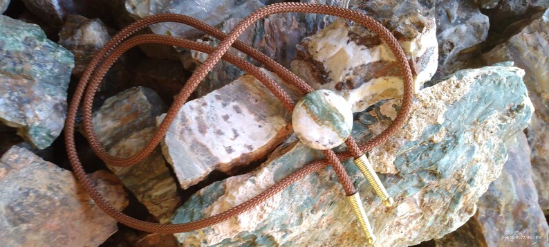 May include: A brown leather bolo tie with a round green and white agate stone pendant. The bolo tie is displayed on a bed of green and white rocks.