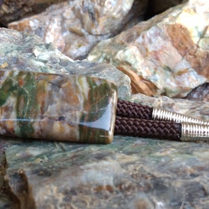 May include: A bolo tie featuring a rectangular stone pendant with a brown, green, and beige pattern. The pendant is attached to a braided brown cord with silver-colored metal tips. The background is a collection of rocks.