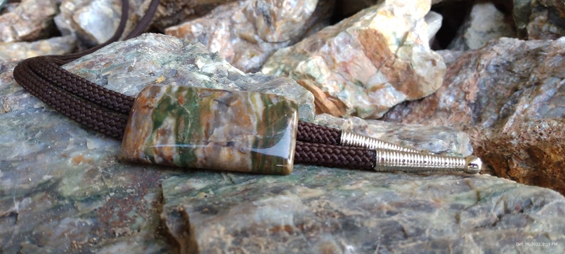 May include: A bolo tie featuring a rectangular stone pendant with a brown, green, and beige pattern. The pendant is attached to a braided brown cord with silver-colored metal tips. The background is a collection of rocks.