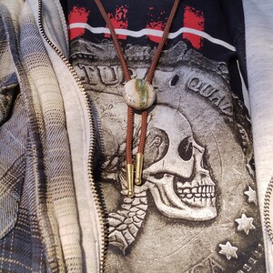 May include: A person wearing a grey hooded sweatshirt over a black t-shirt with a skull graphic and the words "Full of Qual" printed on it. The person is also wearing a brown leather bolo tie with a green and brown stone pendant. The person is wearing a blue and yellow plaid flannel shirt over the sweatshirt.
