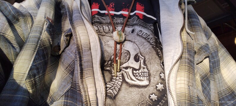 May include: A person wearing a grey hooded sweatshirt over a black t-shirt with a skull graphic and the words "Full of Qual" printed on it. The person is also wearing a brown leather bolo tie with a green and brown stone pendant. The person is wearing a blue and yellow plaid flannel shirt over the sweatshirt.