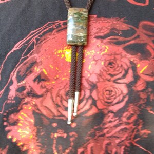May include: A bolo tie with a rectangular, mottled green and brown stone pendant. The cord is dark brown, and the tips are silver. The person is wearing a black t-shirt with a red graphic design.