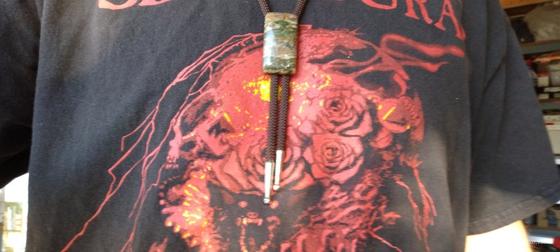 May include: A bolo tie with a rectangular, mottled green and brown stone pendant. The cord is dark brown, and the tips are silver. The person is wearing a black t-shirt with a red graphic design.