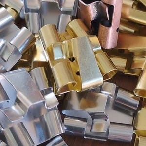 May include: A collection of metal clips in various colors, including silver, gold, and copper. The clips have a unique shape with a curved design and are likely used for fastening or holding items together. The image is a close-up shot.