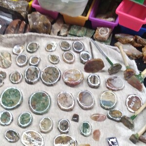 May include: A collection of polished stones and rock specimens, including round and oval shapes with various colors and patterns. Some stones have a white border. Also visible are small tools with wooden handles.