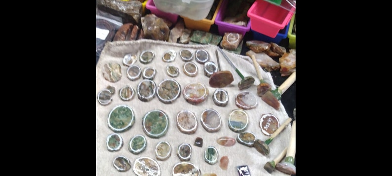 May include: A collection of polished stones and rock specimens, including round and oval shapes with various colors and patterns. Some stones have a white border. Also visible are small tools with wooden handles.