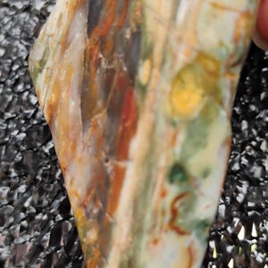 May include: A bolo tie featuring a polished, irregularly shaped stone pendant. The stone displays a mix of colors including orange, red, green, and cream, with a dark brown leather cord.
