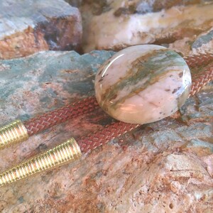 May include: A brown leather bolo tie with a gold-toned metal tip and a round, pink and green agate stone pendant.