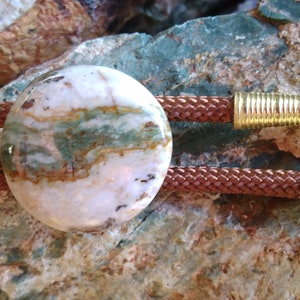 May include: A brown leather bolo tie with a round, white, green, and brown agate stone pendant. The ends of the tie have gold metal tips.