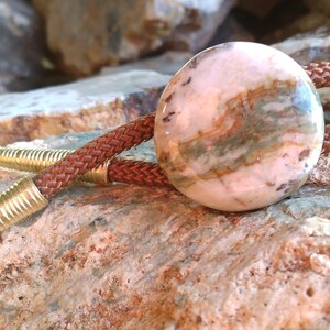 May include: A brown leather bolo tie with a round, pink and brown stone pendant. The stone has a swirling pattern and is set in a gold-toned metal.