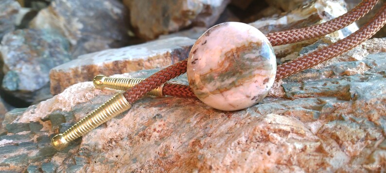 May include: A brown leather bolo tie with a round, pink and brown stone pendant. The stone has a swirling pattern and is set in a gold-toned metal.