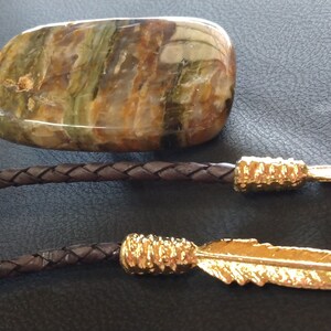 May include: A brown leather bolo tie with a gold-colored arrowhead and feather tip. The bolo tie features a large, flat, green and brown stone pendant.
