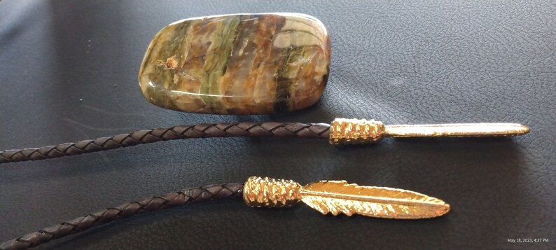 May include: A brown leather bolo tie with a gold-colored arrowhead and feather tip. The bolo tie features a large, flat, green and brown stone pendant.