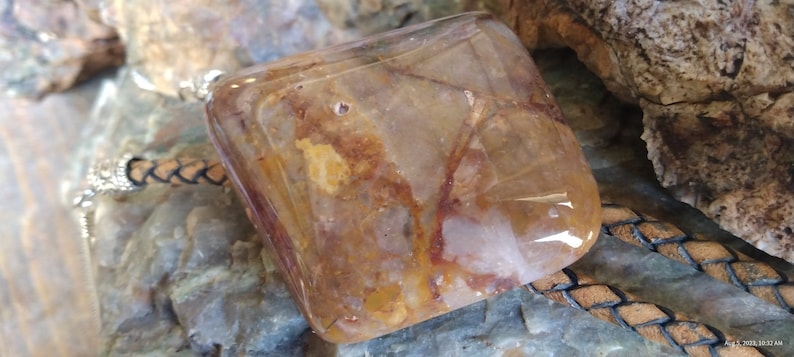 May include: A square-shaped pendant made of clear quartz with brown and white inclusions. The pendant is attached to a brown leather cord.