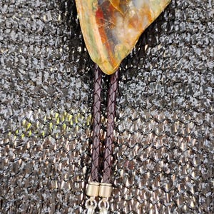 May include: A bolo tie featuring a multi-colored, irregularly shaped stone pendant. The pendant is suspended from a braided brown leather cord, with silver-toned feather-shaped accents and orange-colored stone details.