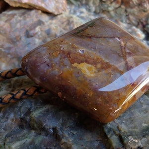 May include: A brown and white agate stone bolo tie with a leather cord. The stone is polished and has a smooth, shiny surface.
