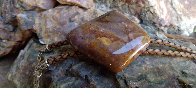 May include: A brown and white agate stone bolo tie with a leather cord. The stone is polished and has a smooth, shiny surface.
