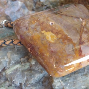 May include: A brown leather bolo tie with a large, clear, square-shaped stone pendant with brown and orange inclusions. The stone is set in a silver bezel.