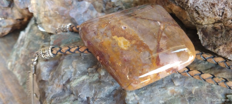 May include: A brown leather bolo tie with a large, clear, square-shaped stone pendant with brown and orange inclusions. The stone is set in a silver bezel.