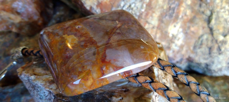 May include: A bolo tie with a large, polished, multi-colored agate stone. The stone has shades of brown, orange, and white. The bolo tie is made of brown leather with a braided design.