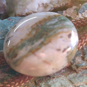 May include: A close-up of a round, smooth, pink and green agate stone with brown and white swirls. The stone is on a brown braided cord.