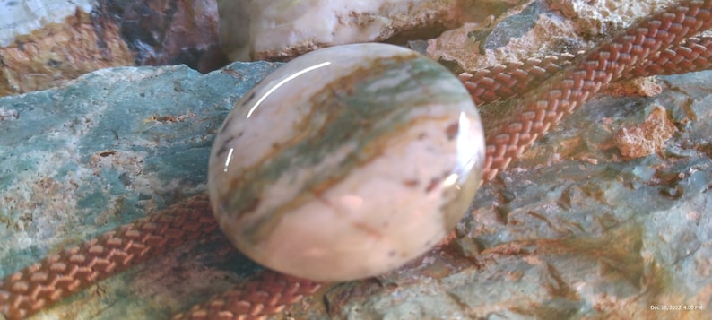 May include: A close-up of a round, smooth, pink and green agate stone with brown and white swirls. The stone is on a brown braided cord.