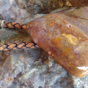 May include: A brown leather bolo tie with a large, irregularly shaped, polished agate stone pendant. The stone has a mix of brown, orange, and white colors.