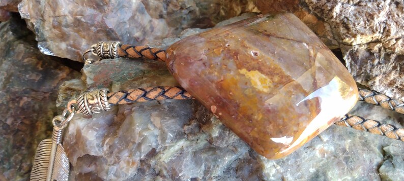 May include: A brown leather bolo tie with a large, irregularly shaped, polished agate stone pendant. The stone has a mix of brown, orange, and white colors.