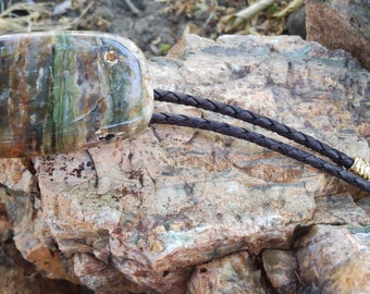 May include: A bolo tie with a large, green, brown, and white agate stone pendant. The bolo tie is made of brown leather cord with gold-colored tips.