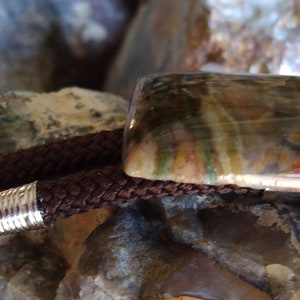 May include: A bolo tie featuring a rectangular, polished stone with brown, green, and red hues. The stone is set on a silver-toned metal base, paired with a braided brown cord and silver tips. A unique accessory.