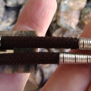 May include: Close-up of a bolo tie. The tie features a dark brown braided cord with silver-colored metal tips and a decorative slide. The slide is rectangular with a mottled green and brown pattern.