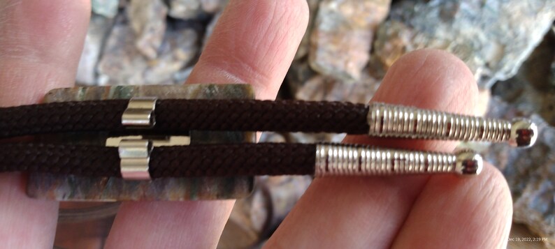 May include: Close-up of a bolo tie. The tie features a dark brown braided cord with silver-colored metal tips and a decorative slide. The slide is rectangular with a mottled green and brown pattern.