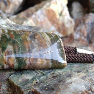 May include: A rectangular bolo tie with a polished stone centerpiece. The stone features a mix of green, brown, and tan colors. The tie has a brown braided cord and silver-colored metal tips.