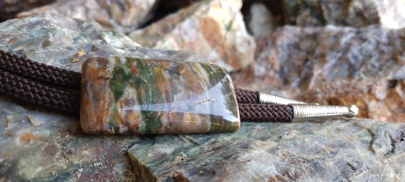 May include: A rectangular bolo tie with a polished stone centerpiece. The stone features a mix of green, brown, and tan colors. The tie has a brown braided cord and silver-colored metal tips.
