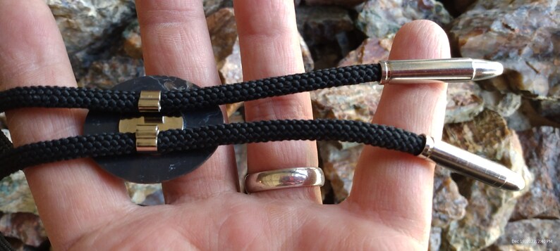Pu&ograve; includere: Un bolo tie nero con uno slider rotondo in metallo nero e due punte a proiettile argentate.