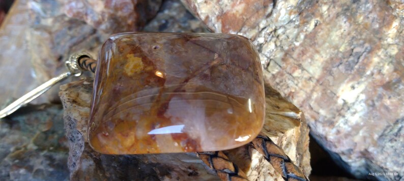 May include: A square-shaped, translucent, brown and white agate stone with a polished surface. The stone is set in a leather cord.