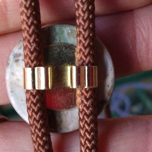 May include: A brown leather bolo tie with a round, multi-colored stone pendant. The stone has a white, green, and red pattern. The tie is secured with gold metal clasps.