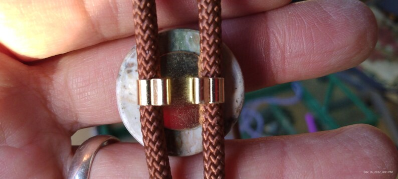 May include: A brown leather bolo tie with a round, multi-colored stone pendant. The stone has a white, green, and red pattern. The tie is secured with gold metal clasps.