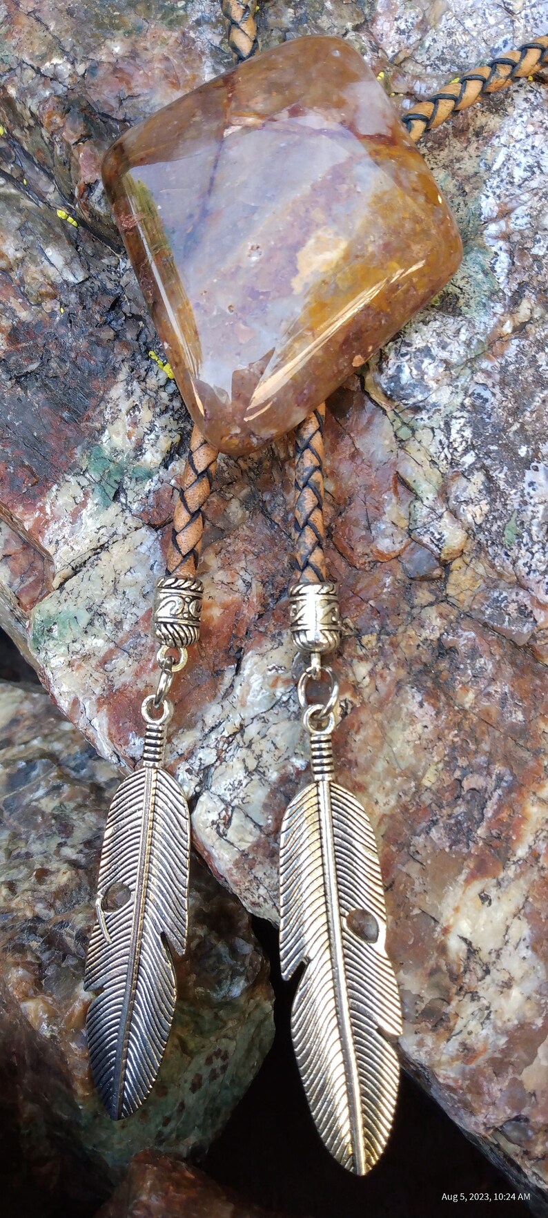 May include: A brown leather bolo tie with a large, clear, brown and yellow agate stone pendant. The tie has silver feather charms hanging from the ends.