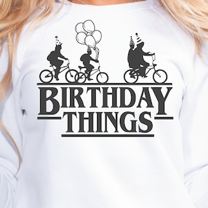 Pode incluir: Camisola branca com um gr&aacute;fico preto de tr&ecirc;s pessoas a andar de bicicleta com chap&eacute;us de festa. O texto "Birthday Things" est&aacute; abaixo do gr&aacute;fico.