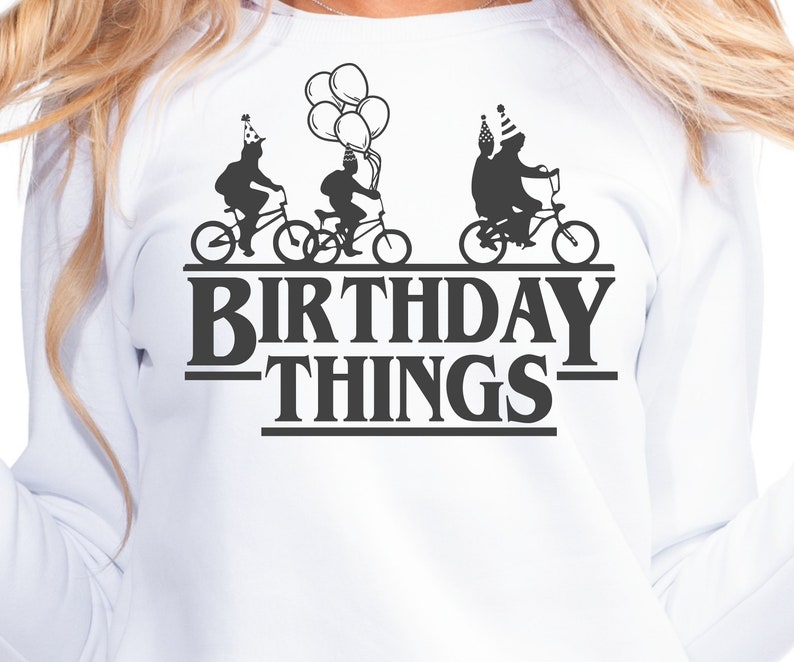 Pode incluir: Camisola branca com um gr&aacute;fico preto de tr&ecirc;s pessoas a andar de bicicleta com chap&eacute;us de festa. O texto "Birthday Things" est&aacute; abaixo do gr&aacute;fico.
