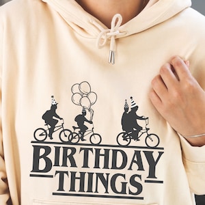 Pode incluir: Um moletom bege com um gr&aacute;fico preto de tr&ecirc;s figuras andando de bicicleta com bal&otilde;es e o texto "Birthday Things".