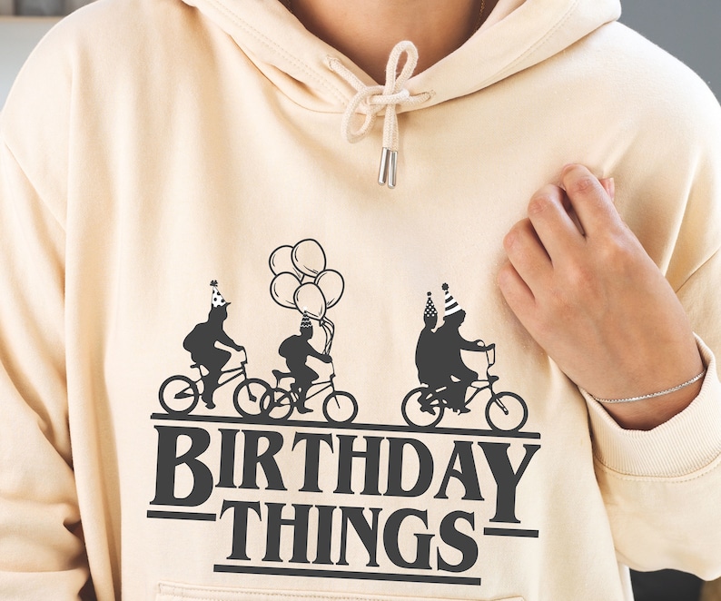 Pode incluir: Um moletom bege com um gr&aacute;fico preto de tr&ecirc;s figuras andando de bicicleta com bal&otilde;es e o texto "Birthday Things".