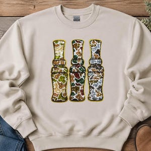 May include: A beige crewneck sweatshirt featuring a graphic of three vertical duck calls. Each call is filled with a different camouflage pattern rendered in shimmering sequins, outlined in gold thread against the light fabric background.