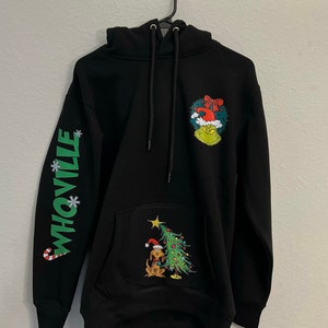 May include: Black hooded sweatshirt with a green Grinch character wearing a Santa hat and a Christmas tree with a dog wearing a Santa hat on the front. The left sleeve has the word "Whoville" in green with snowflakes.