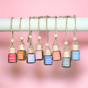 May include: Eight glass bottles with wooden tops and string hangers, hanging from a white bar. Each bottle contains a scented oil with a different label, including "Strawberry", "Blood Orange", "Cotton Candy", "Birthday Cake", "Fruity Cereal", "Strawberry Cake", "Tistaeftio And Coennid", and "Linea".