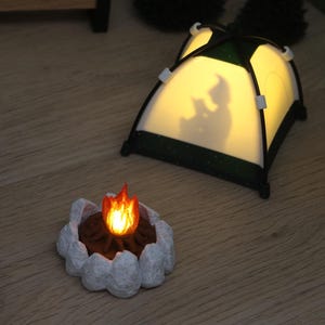 Könnte beinhalten: Eine Miniatur-Camping-Szene mit einem beleuchteten Zelt, das die Silhouette einer lesenden Figur zeigt, einem kleinen Lagerfeuer mit orangefarbenen Flammen, einer Holzbank mit der Aufschrift Tomte, einem Holztisch mit Kompass und zwei kleinen Tannen.